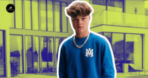 A Man In A Blue Sweater Stands In Front Of A Building, Looking Towards The Camera With A Neutral Expression.