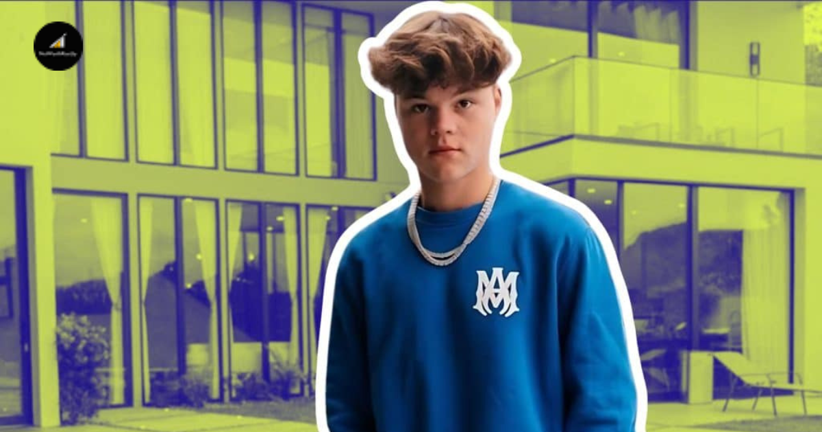 A Man In A Blue Sweater Stands In Front Of A Building, Looking Towards The Camera With A Neutral Expression.